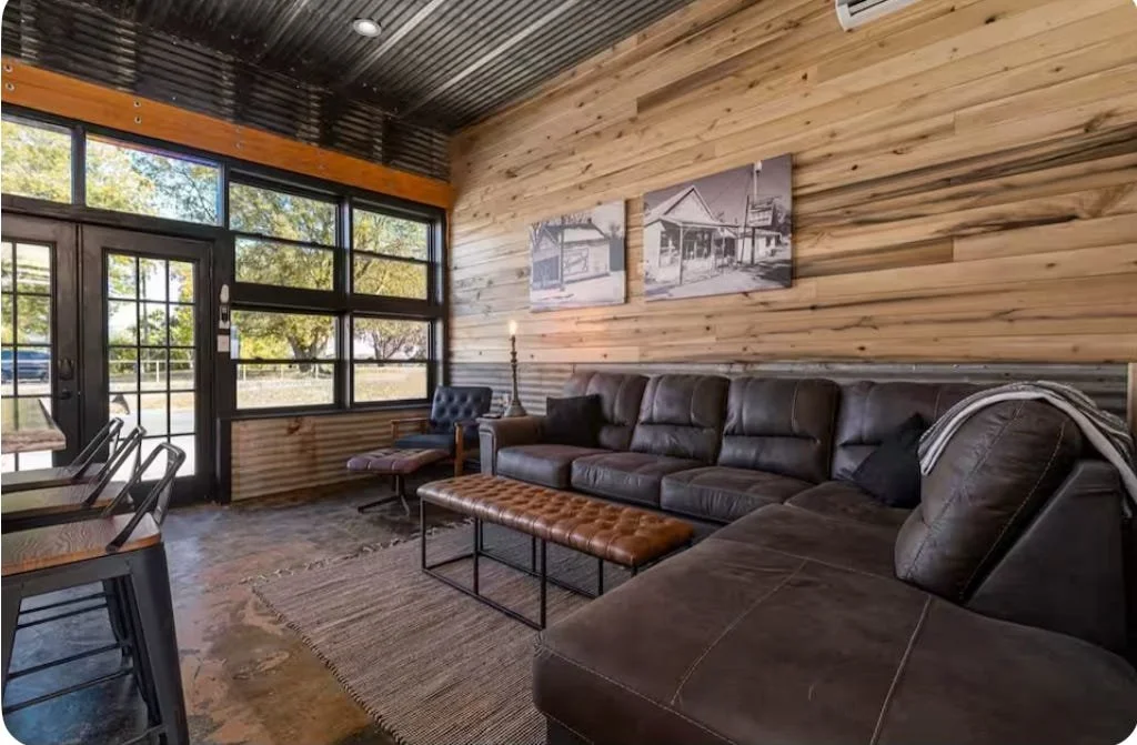 Interior of a rustic-style lounge area with a leather sofa, armchair, wooden wall paneling, large windows, and minimal wall art.