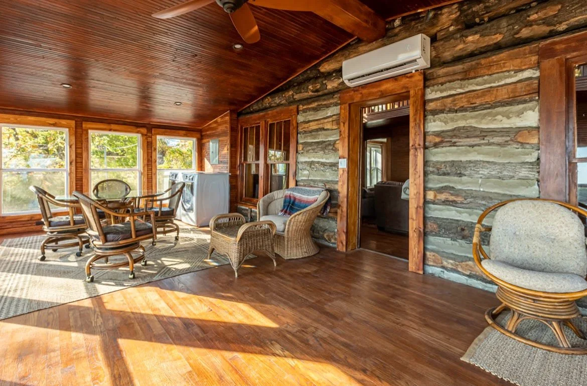 A cozy indoor room with wooden walls, ceiling, and floor. It features multiple windows, with sunlight streaming in. The room contains dining chairs around a round glass table, a wicker armchair with a cushion, a rocking chair with a blanket, and an a