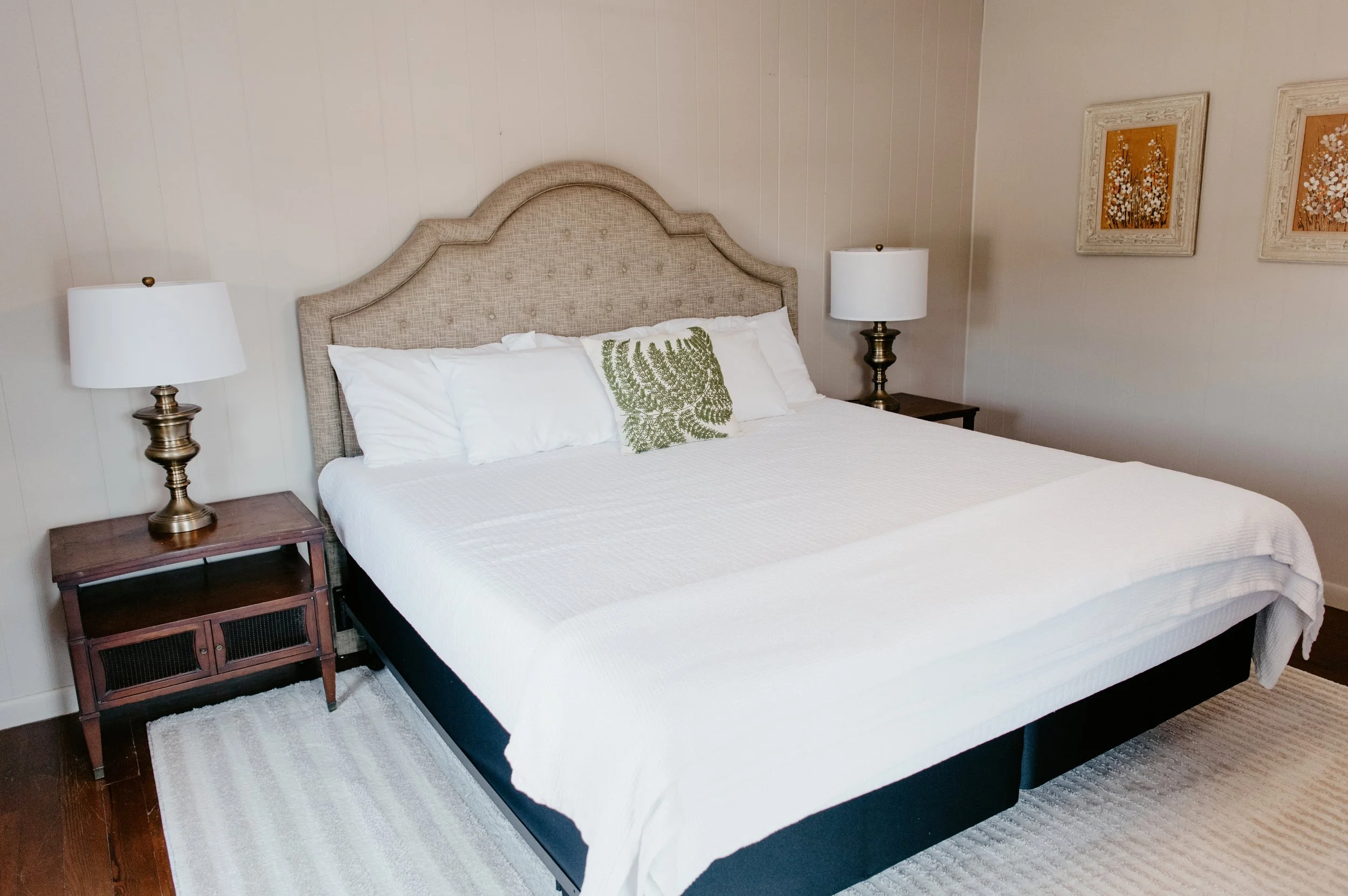 A neatly made bed with white bedding, decorative pillows, and an upholstered beige headboard in a cozy bedroom. There are two nightstands with lamps on each side of the bed, and artwork hanging on the wall.