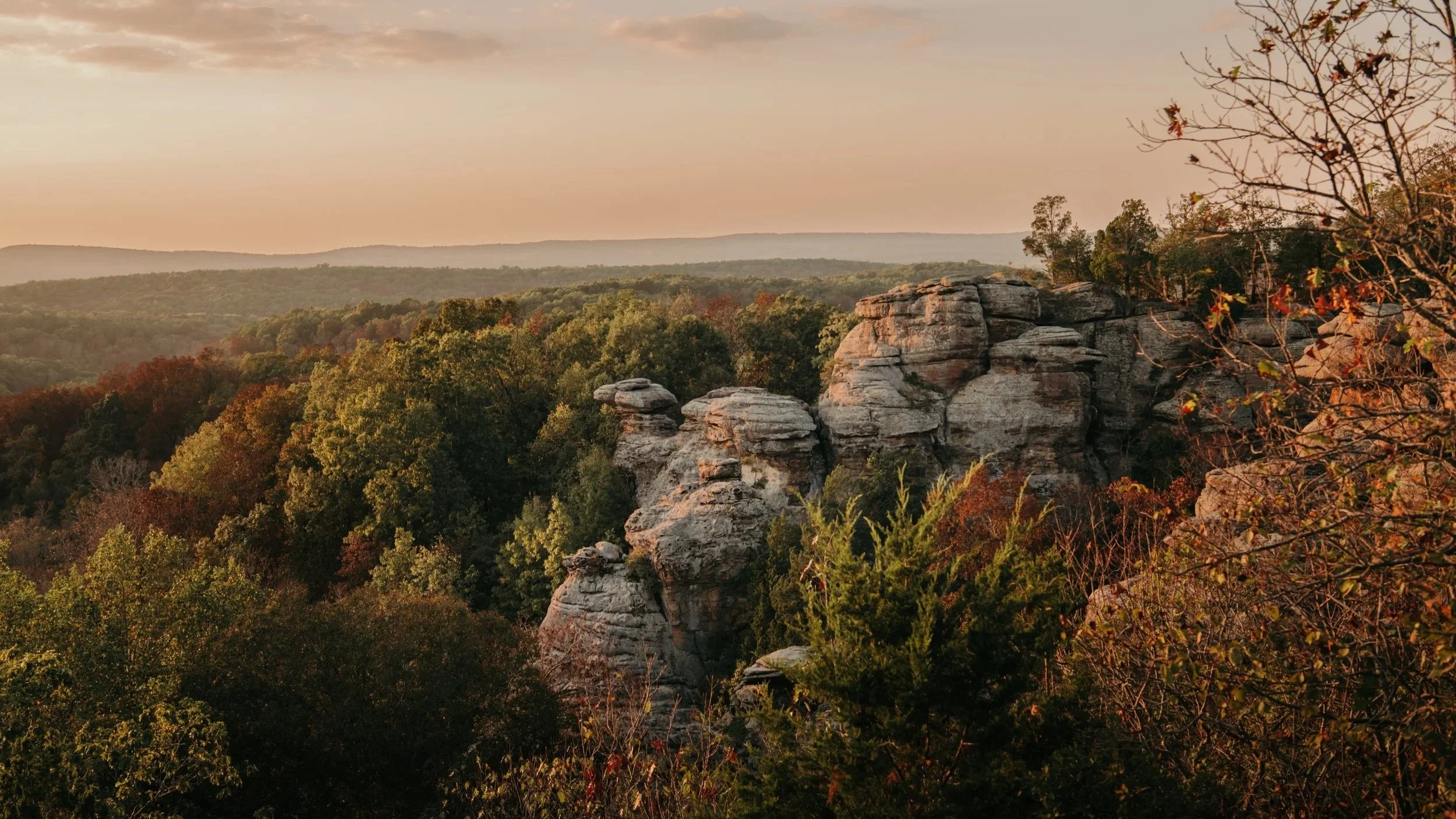 Sunset view of a rocky cliff surrounded by dense forest with rolling hills in the background.