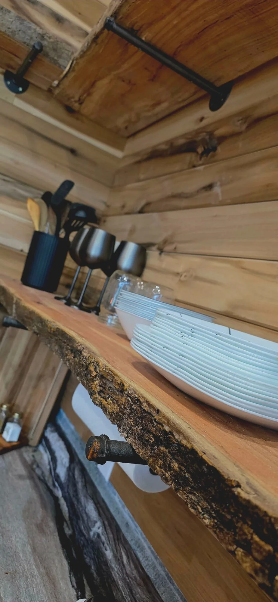 Rustic kitchen shelf with white plates, glassware, and black cups, mounted on wooden walls.