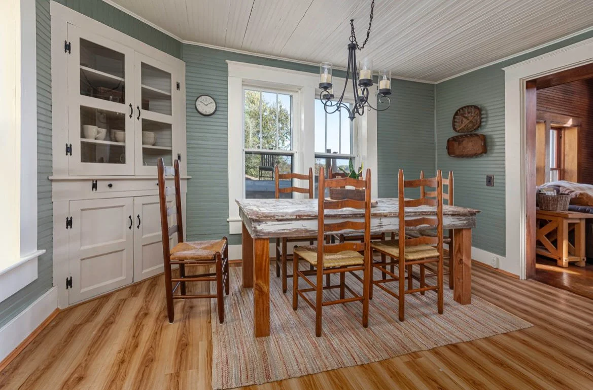 A dining room with a rustic wooden table, six wooden chairs, a white china cabinet, a colorful rug, a window, and a chandelier, with a view into a living space with wooden accents.