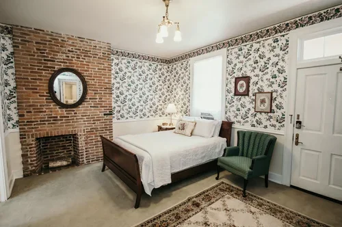 Cozy bedroom with floral wallpaper, a white bed, green armchair, brick fireplace, and a window with natural light.