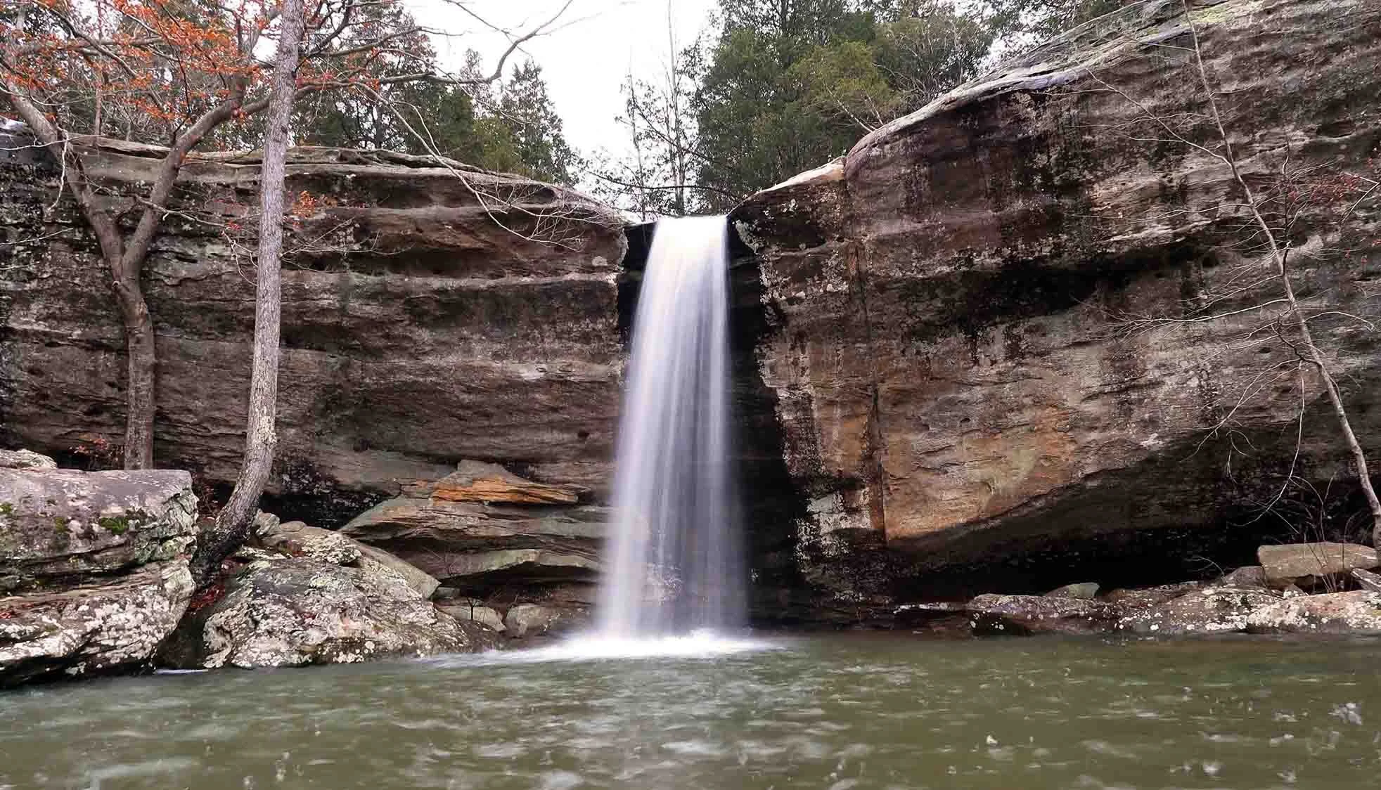 A small waterfall cascading down a rocky cliff surrounded by trees and water at the base.