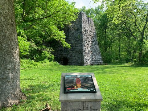 A historical stone structure in a forested area with an informational plaque in the foreground.