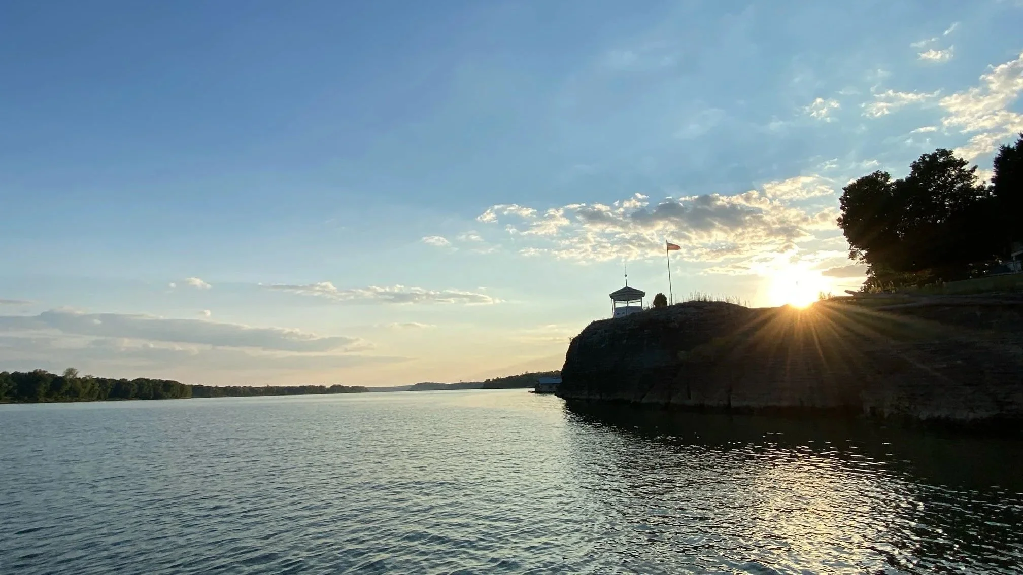 Sunset over a river with a small building and American flag on a cliff, trees on the right side, and partly cloudy sky.
