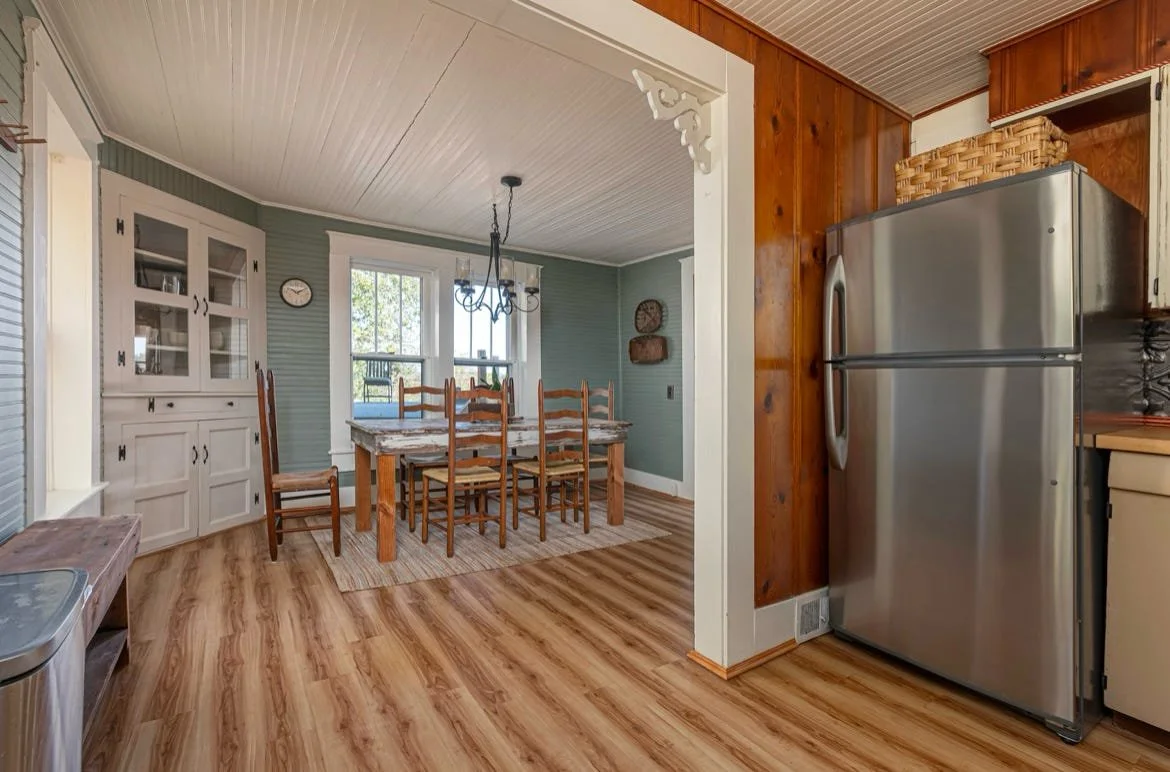 A kitchen opening into a dining area with a wooden dining table and six chairs, a white china cabinet, a window with a view of trees, and a chandelier. The kitchen features a stainless steel refrigerator, wooden wall paneling, and laminate wood floor