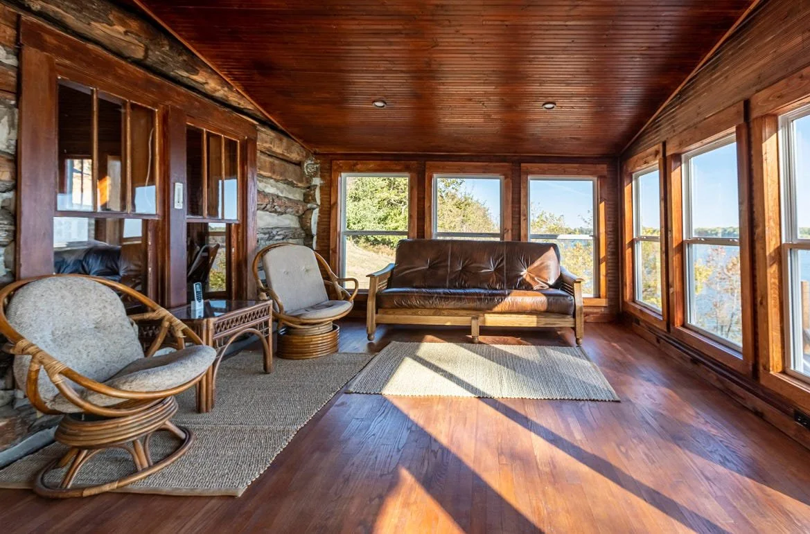 Sunlit enclosed porch with wooden floors, walls, and ceiling, furnished with a leather sofa, bamboo chairs, and area rugs, overlooking trees and a body of water outside.