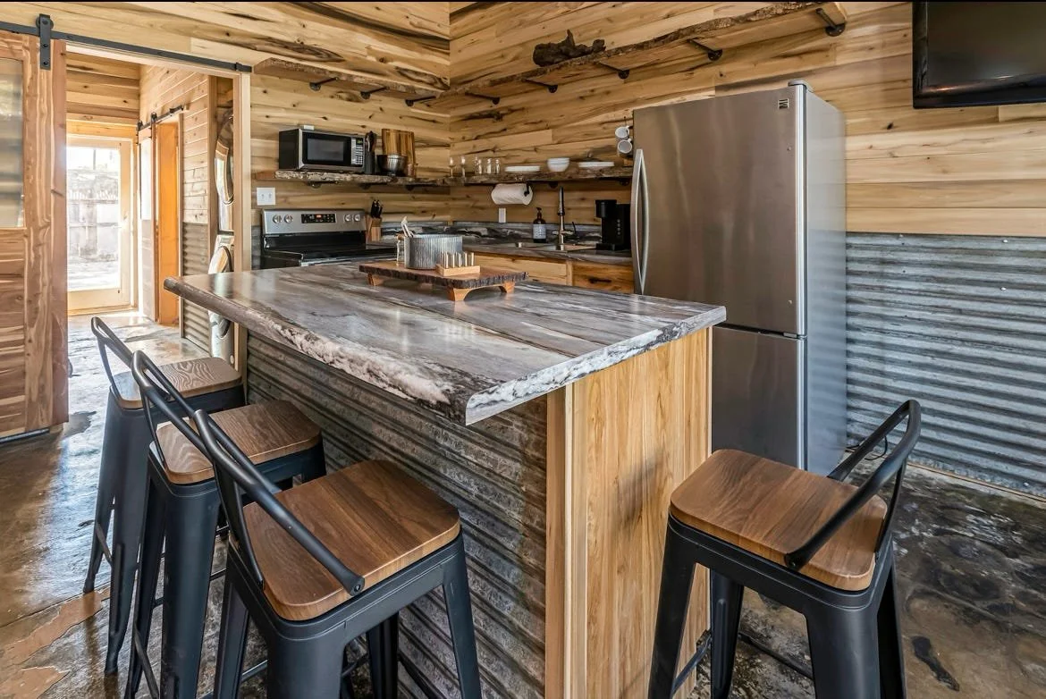 Modern kitchen with wood-paneled walls, a large marble countertop island, and black metal barstools with wooden seats.