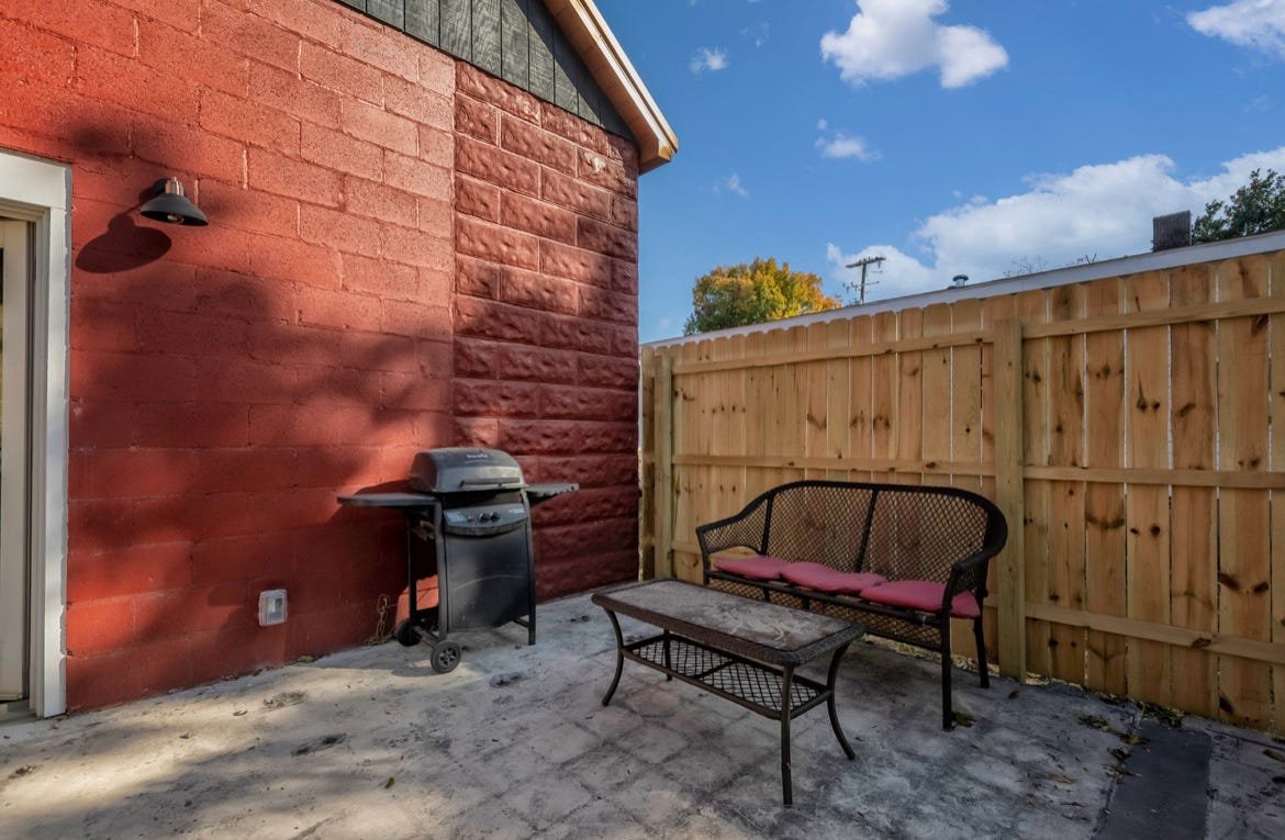 Small outdoor patio area with a brick red house wall, a black barbecue grill, a black metal bench with pink cushions, a small table, and a wooden privacy fence, under a partly cloudy sky.