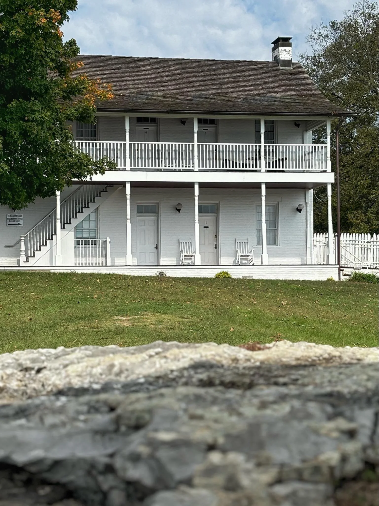 A two-story house with a brown shingle roof, white exterior, and a large front porch with white railings and stairs. There are two rocking chairs on the porch, and the house has multiple windows and a chimney. The house is surrounded by green grass a