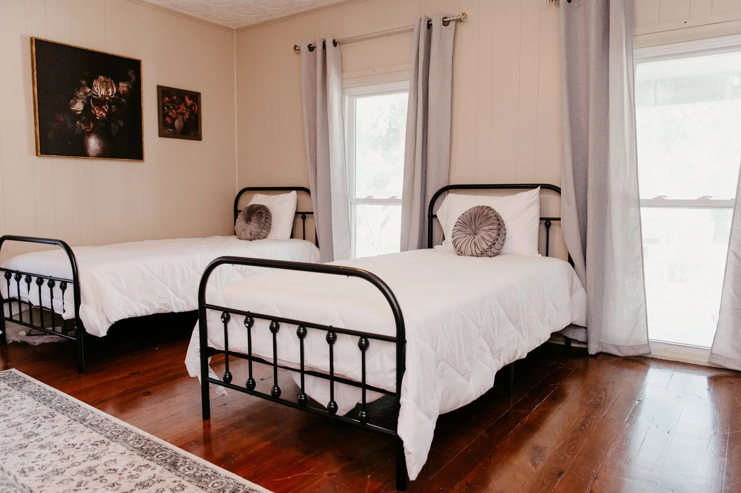 A bedroom with two twin beds with black metal frames, white duvets, and round decorative pillows. There are two windows with light-colored curtains, and framed floral artwork on the wall.