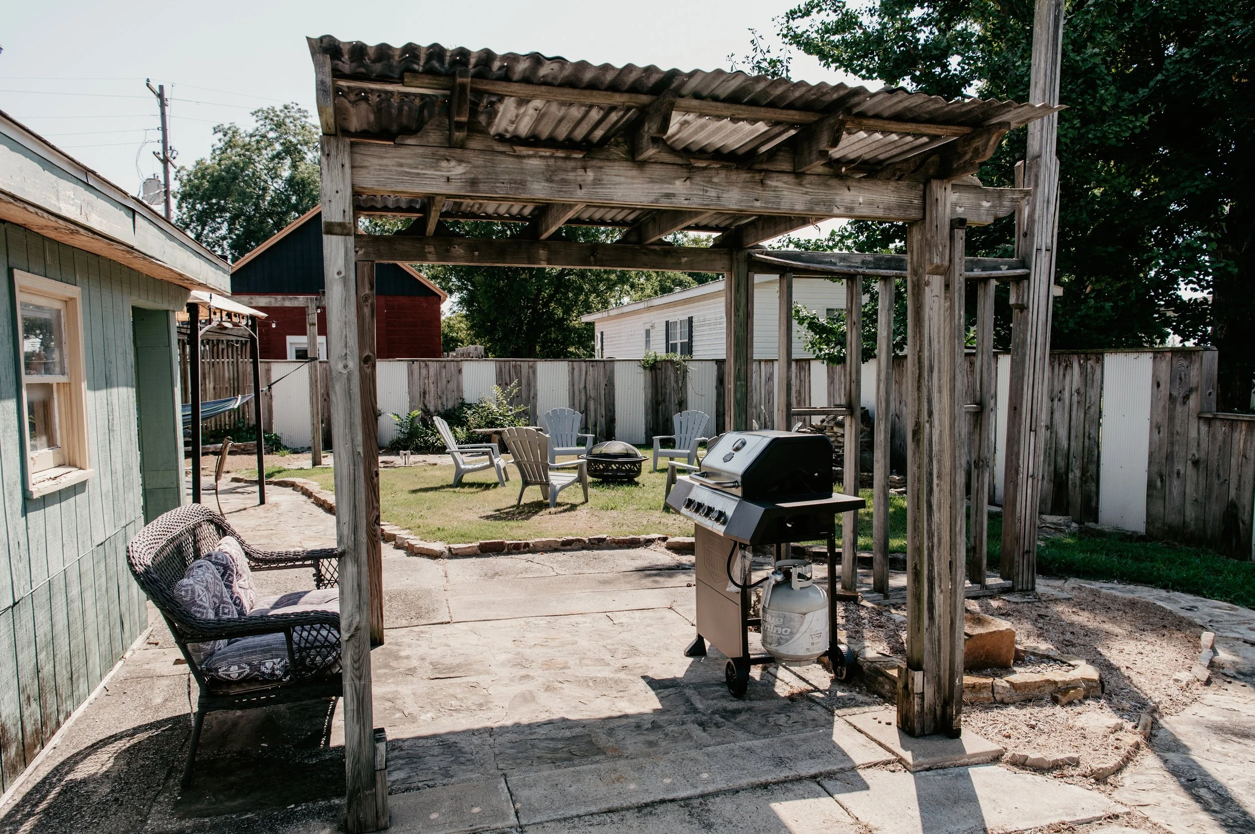 Backyard with a wooden pergola, green lawn, outdoor chairs, barbecue grill, and surrounding houses and trees.