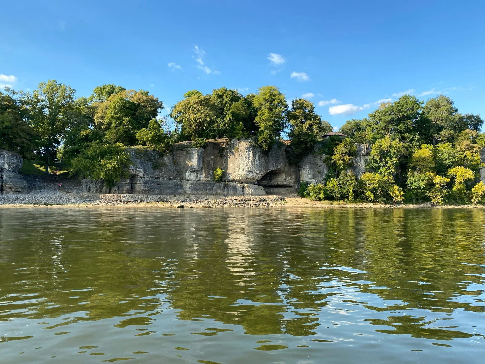 Calm river in the foreground with a rocky shoreline, lush green trees on cliffs in the background, and a bright blue sky with scattered white clouds.