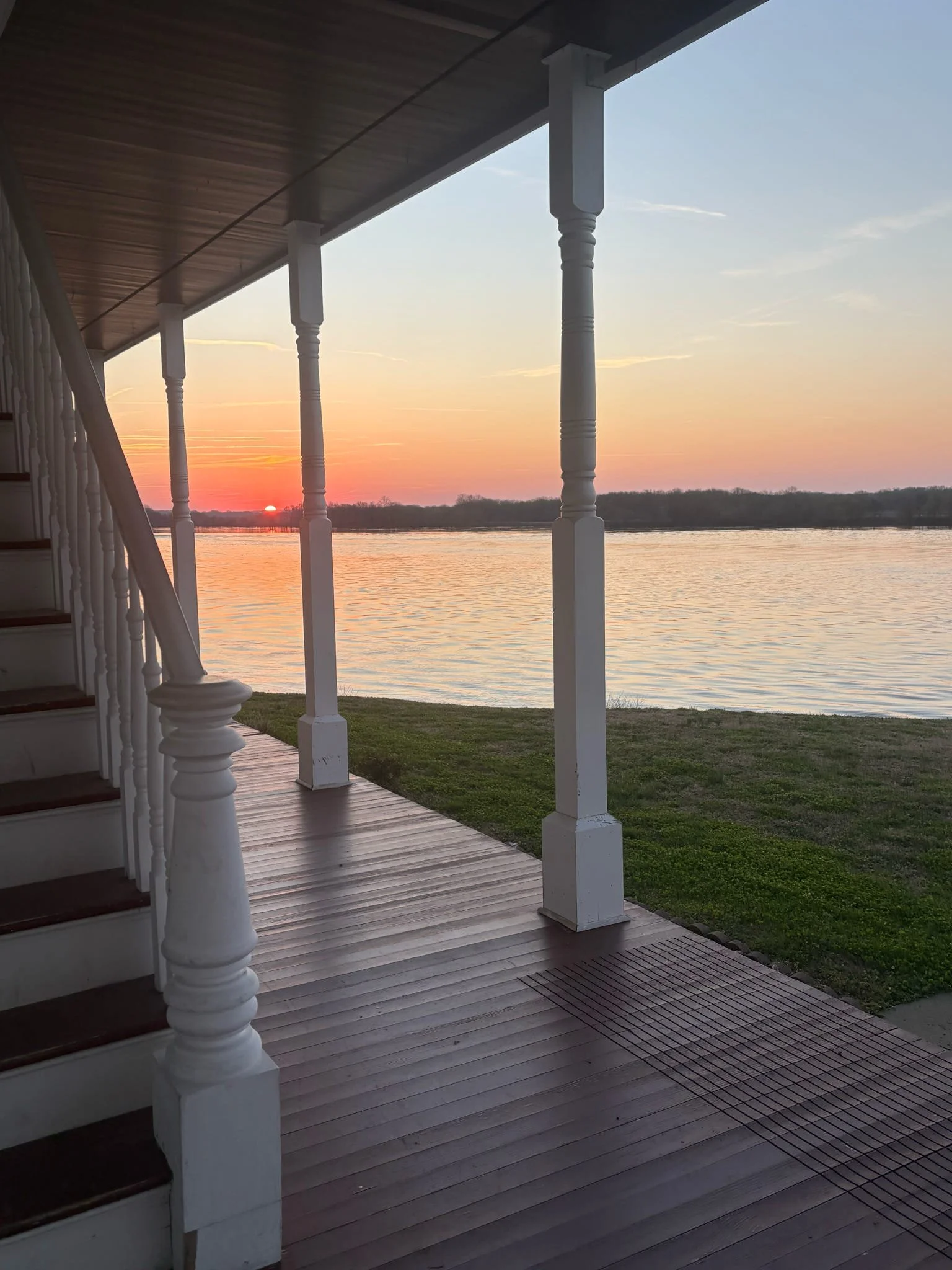 View from a porch overlooking a river at sunset, with the sun near the horizon and colorful sky, supported by white wooden pillars, with stairs visible on the left.