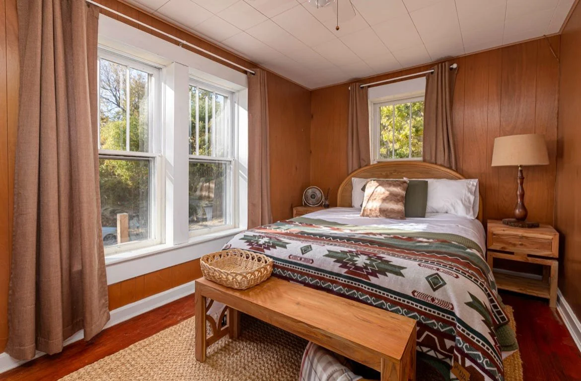 A cozy bedroom with wooden panel walls and large windows showing trees outside. The bed has a patterned bedspread with pillows, a wooden headboard, a nightstand with a lamp, and a small fan. A wooden bench with a basket is at the foot of the bed.