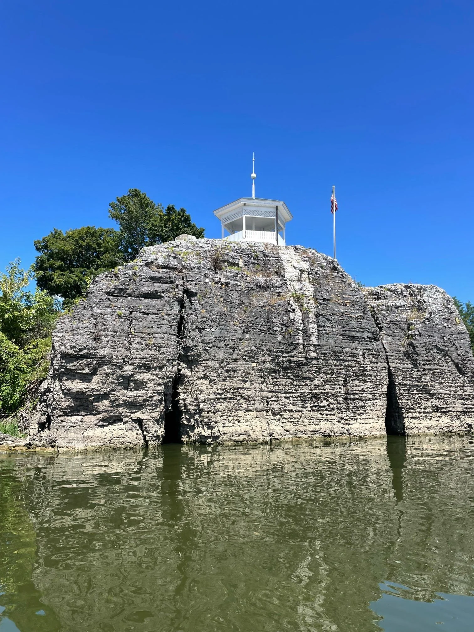 A rocky cliff with a small white pavilion and flagpole on top, overlooking a body of water, trees, and a clear blue sky.
