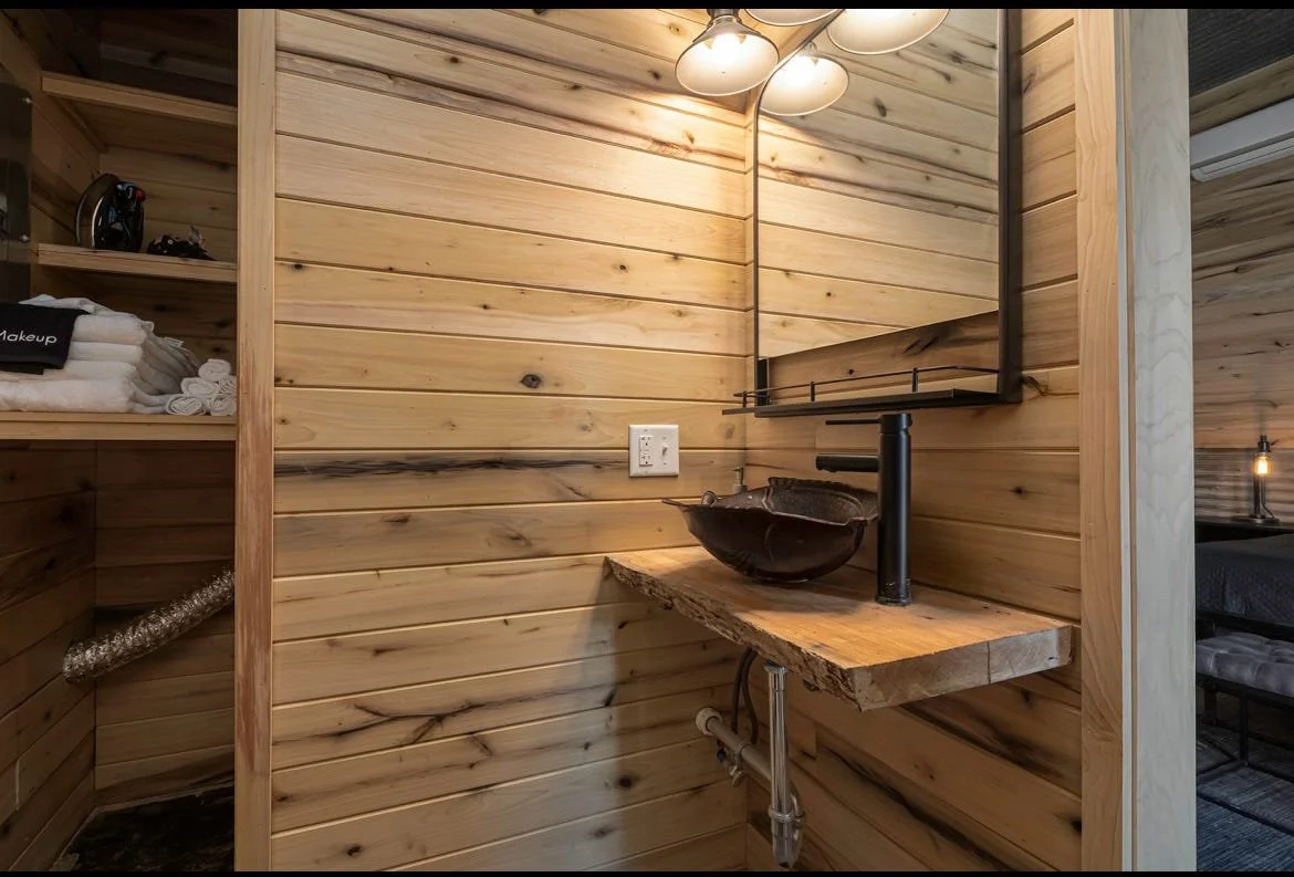 A rustic wooden bathroom vanity with a black vessel sink and a tall, black faucet, set against wood-paneled walls, with above lighting and a mirror.