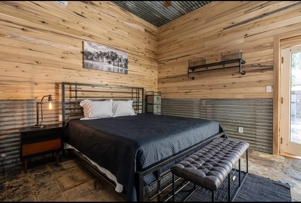 Bedroom with wood-paneled walls, modern black metal bed frame, black quilt, white pillows, bedside table with lamp, wall-mounted shelf, black and white artwork, door to outside.