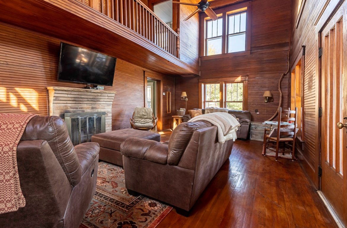 Living room with wood-paneled walls, large windows, and a high ceiling with a ceiling fan. There are leather sofas, a rug, a TV mounted above a brick fireplace, and various chairs and lamps.