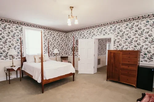 Bedroom with floral wallpaper, wooden bed with white bedding, wooden dresser, nightstand with lamp, and open door leading to another room.