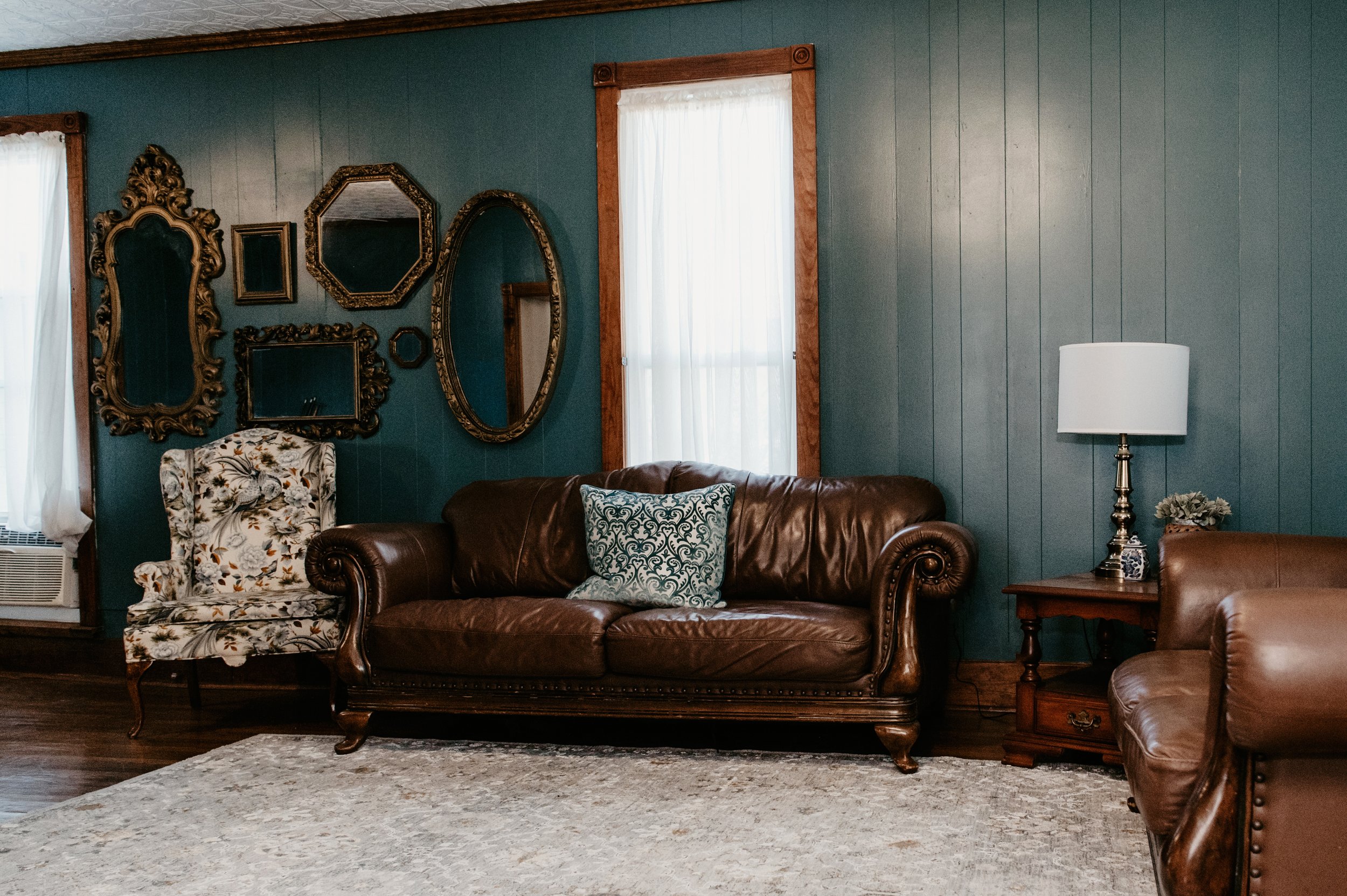 Living room with green paneled walls, decorated with vintage mirrors, a floral armchair, a brown leather sofa with patterned pillows, a wooden side table with a white lamp, and a window with white curtains.