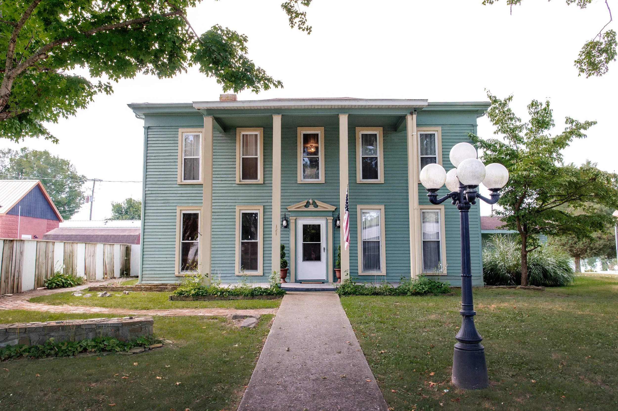 A two-story mint green house with white trim, surrounded by green trees, with a sidewalk and vintage street lamp in front.