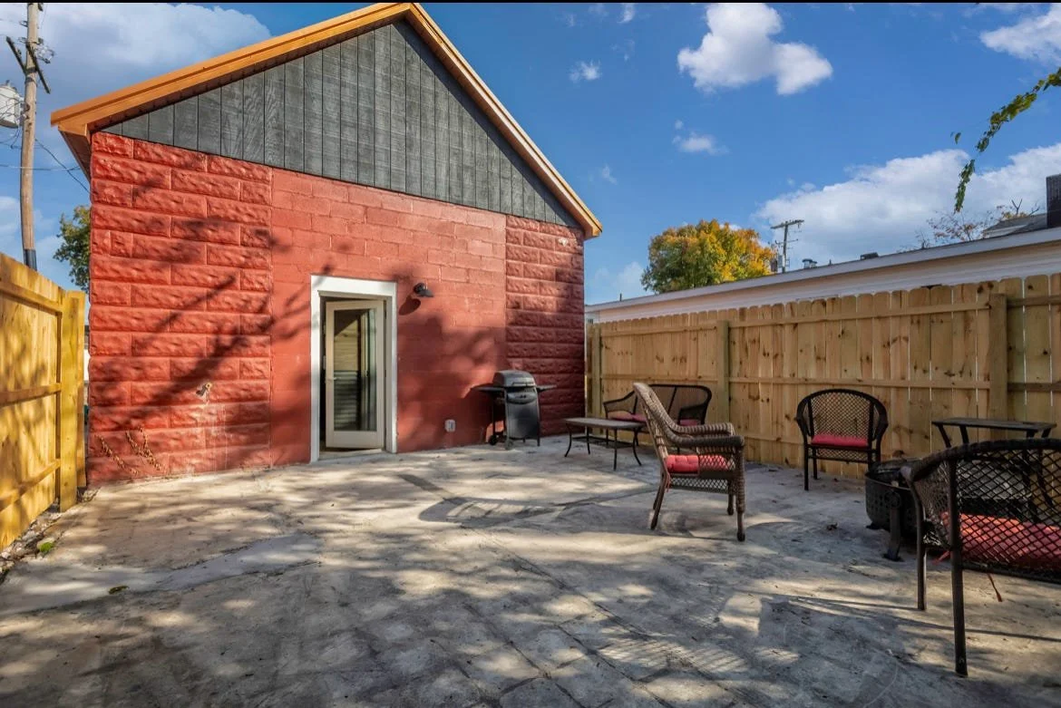 A backyard patio with outdoor furniture, a grill, and a red brick building with a door. The yard is enclosed by a wooden fence, with some shadows from nearby trees.