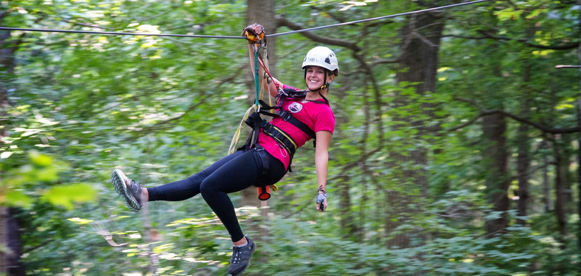 A woman smiling while zip-lining through a dense green forest, wearing a helmet and safety harness.