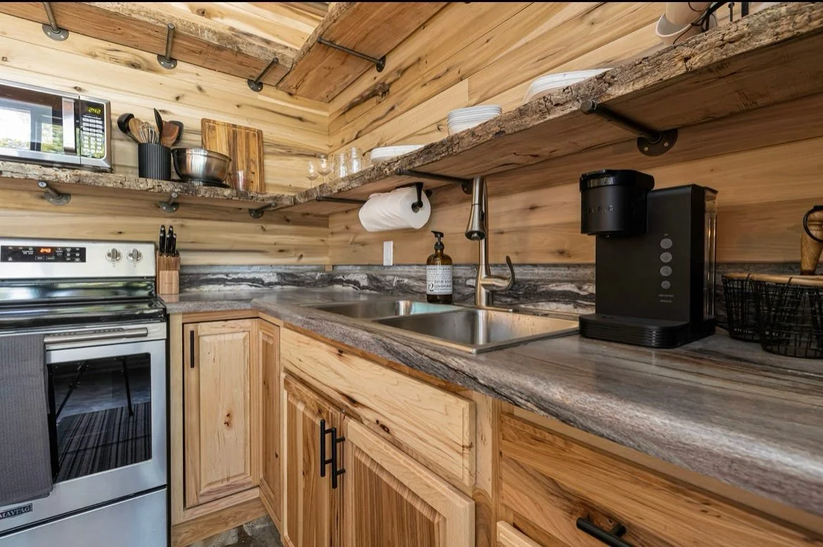A rustic kitchen with wooden cabinets, open wooden shelves, a granite countertop, a stainless steel sink, a black coffee maker, a microwave, and a stove, all set against wood-paneled walls and ceiling.