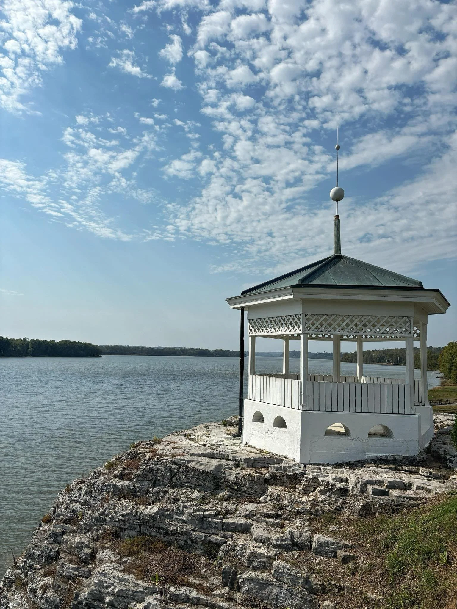 A white gazebo with a metal roof and a tall spire with spherical ornaments, situated on a rocky shoreline next to a body of water, under a blue sky with scattered clouds.