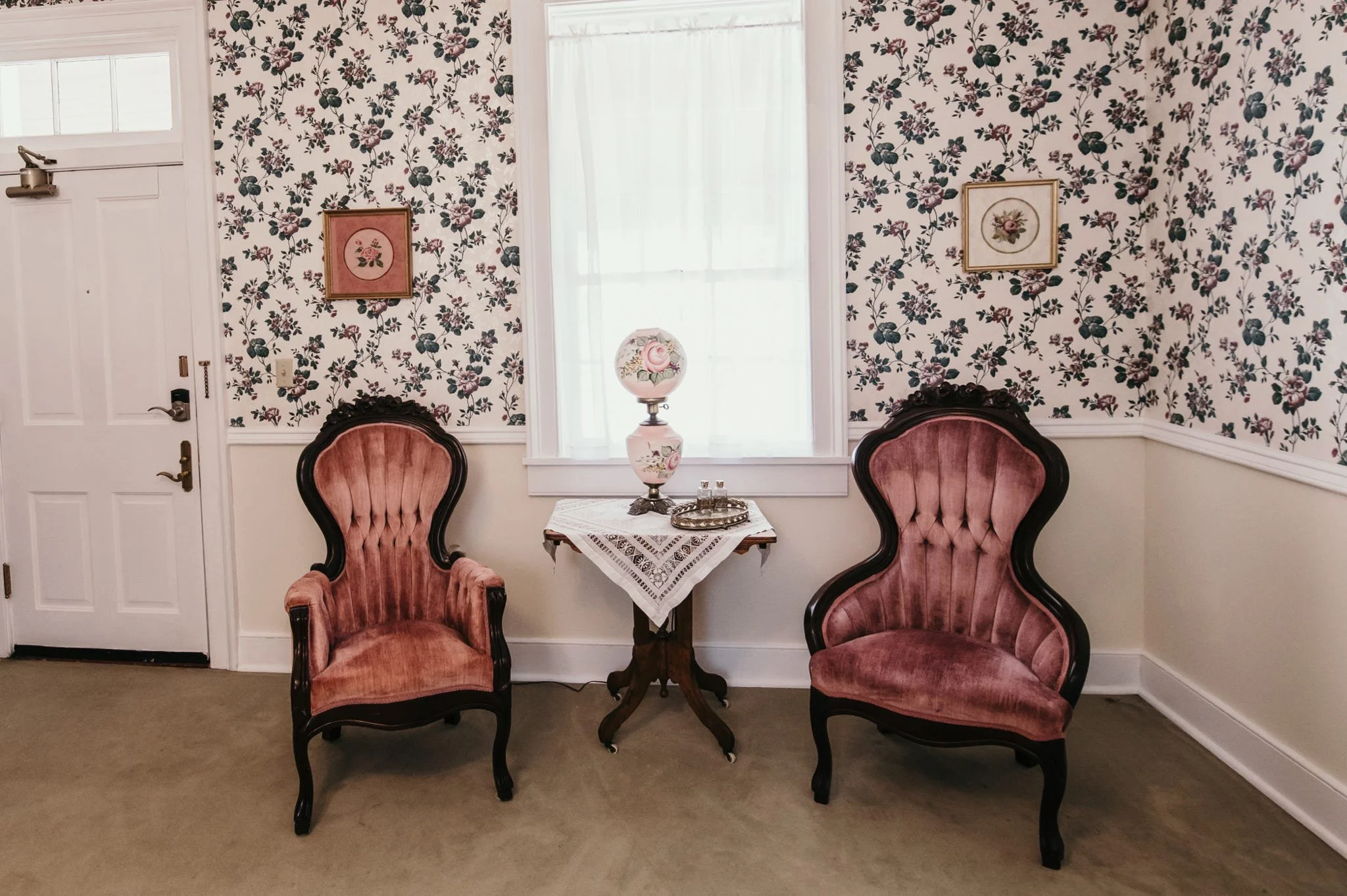 A cozy corner with two vintage pink velvet armchairs and a small wooden table between them. The table has a lace tablecloth, a decorative table lamp with floral design, and a tray. They are in front of a wall covered with floral wallpaper, and a wind