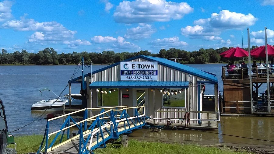 Floating restaurant called E-Town River Restaurant on a river with a ramp leading to the entrance, boats docked nearby, and a deck with red umbrellas and outdoor seating