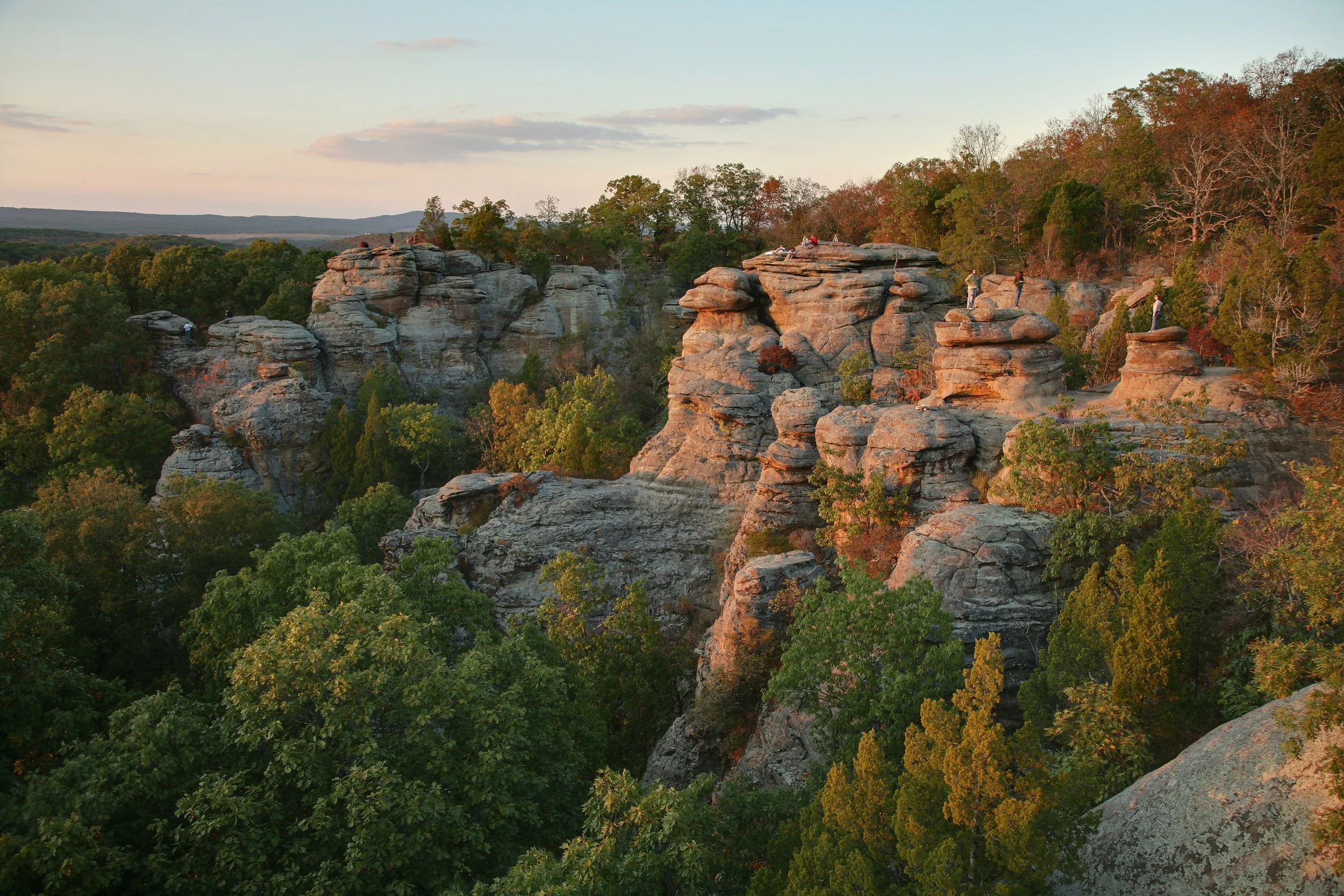 Rock formations with people sitting and climbing, surrounded by trees with autumn foliage, under a partly cloudy sky.