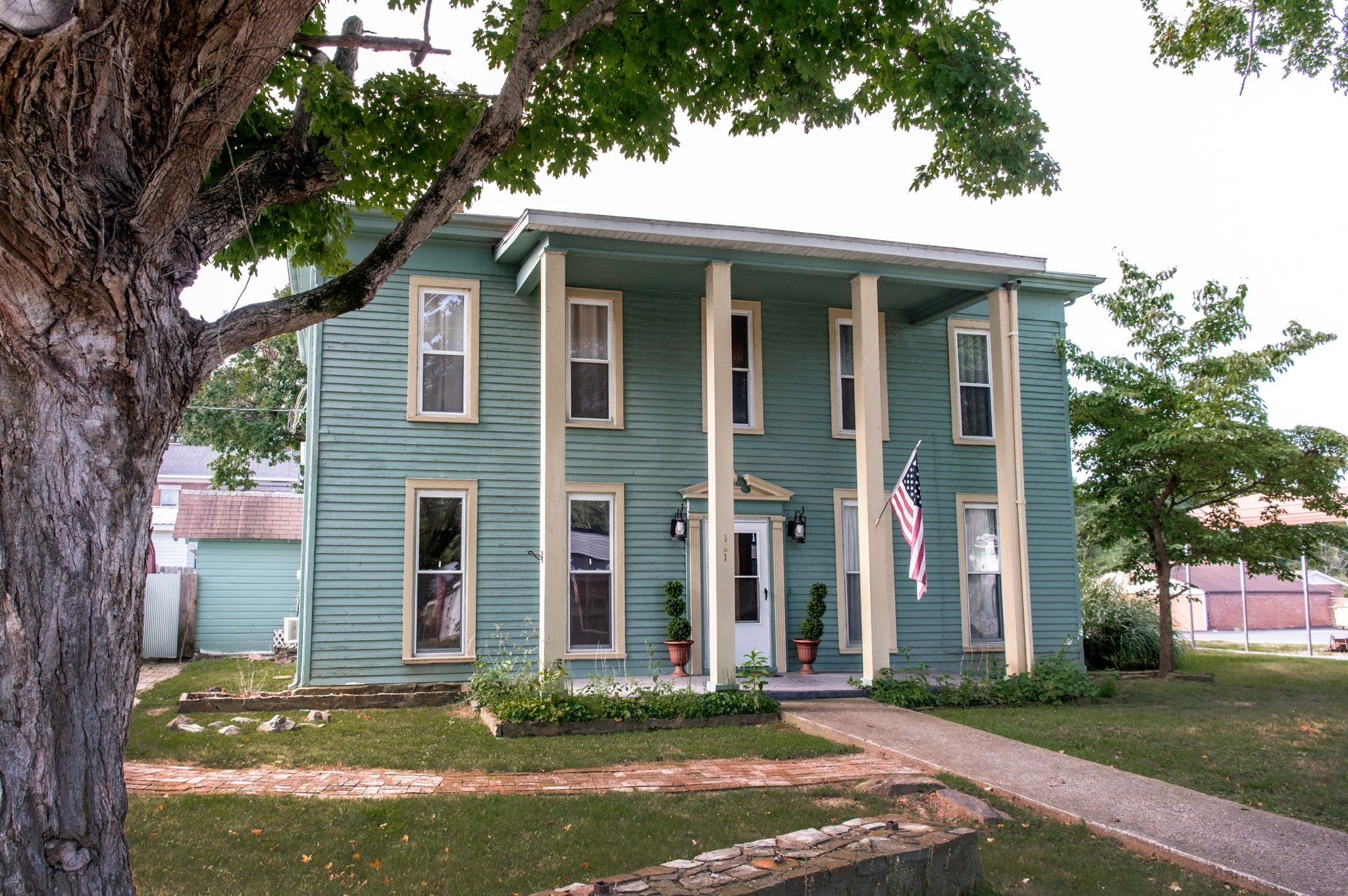 Front of a green house with four large windows on the front, an American flag hanging from the porch, and potted plants at the entrance. A large tree with textured bark and green leaves is in the foreground.