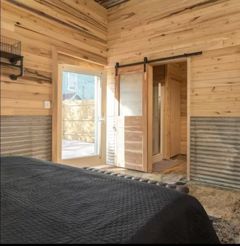 Interior view of a bedroom with wooden panel walls, a sliding barn door, and a glass door leading outside. A bed with a dark quilt is partially visible in the foreground.