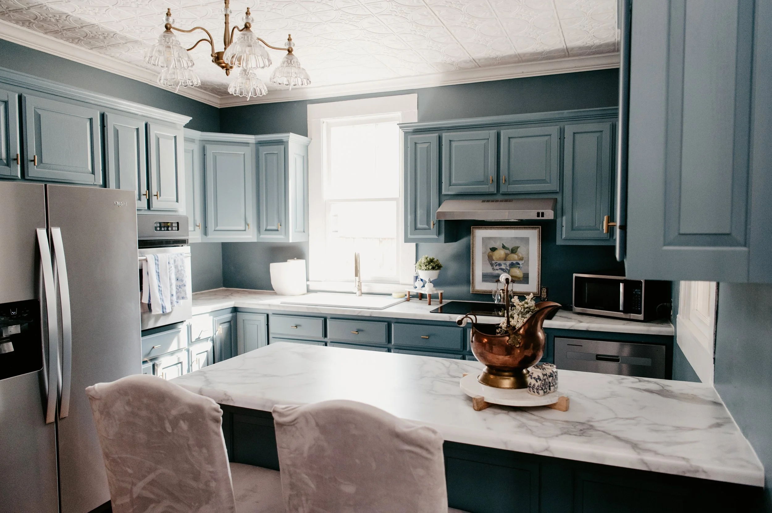 A kitchen with blue cabinets, white marble countertops, a stainless steel refrigerator, a microwave, and a window allowing natural light. There are decorative items on the counter, including a framed fruit artwork, a potted plant, and a copper pitcher on a marble-topped island.