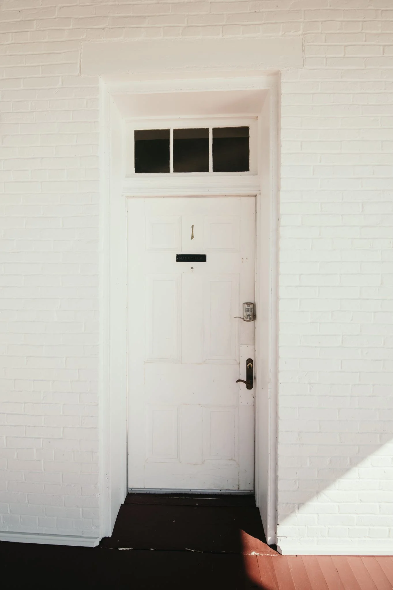 White door with glass panels at the top, set in a white brick wall, with a black mail slot and door handle. The door has the number 1 on it and a black sign beneath with text that isn't legible. There is a dark doormat in front of the door, and sunli