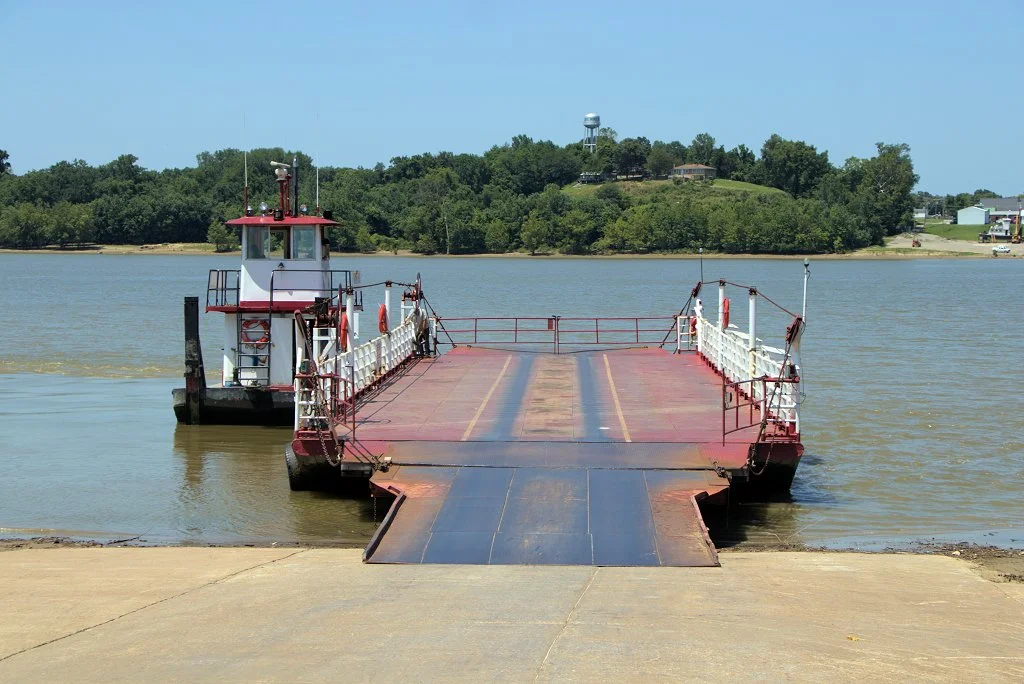 A ferry boat preparing to cross a river with a landmass and trees in the background.