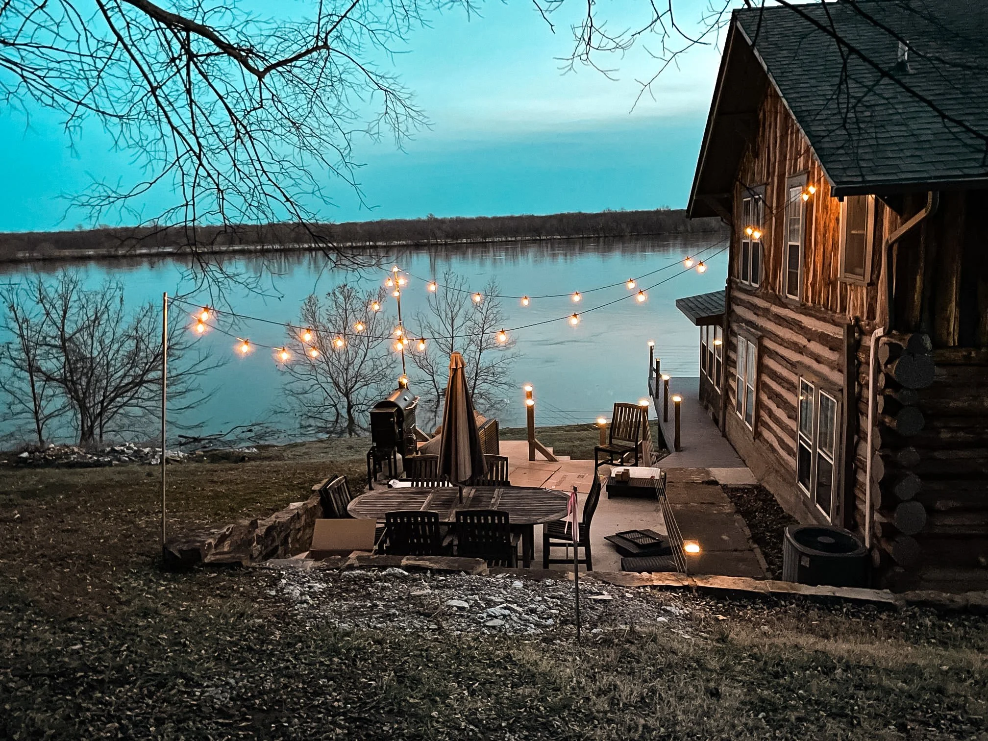 A lakeside backyard scene at dusk with a wooden house featuring string lights, outdoor seating, leafless trees, and a calm lake in the background.