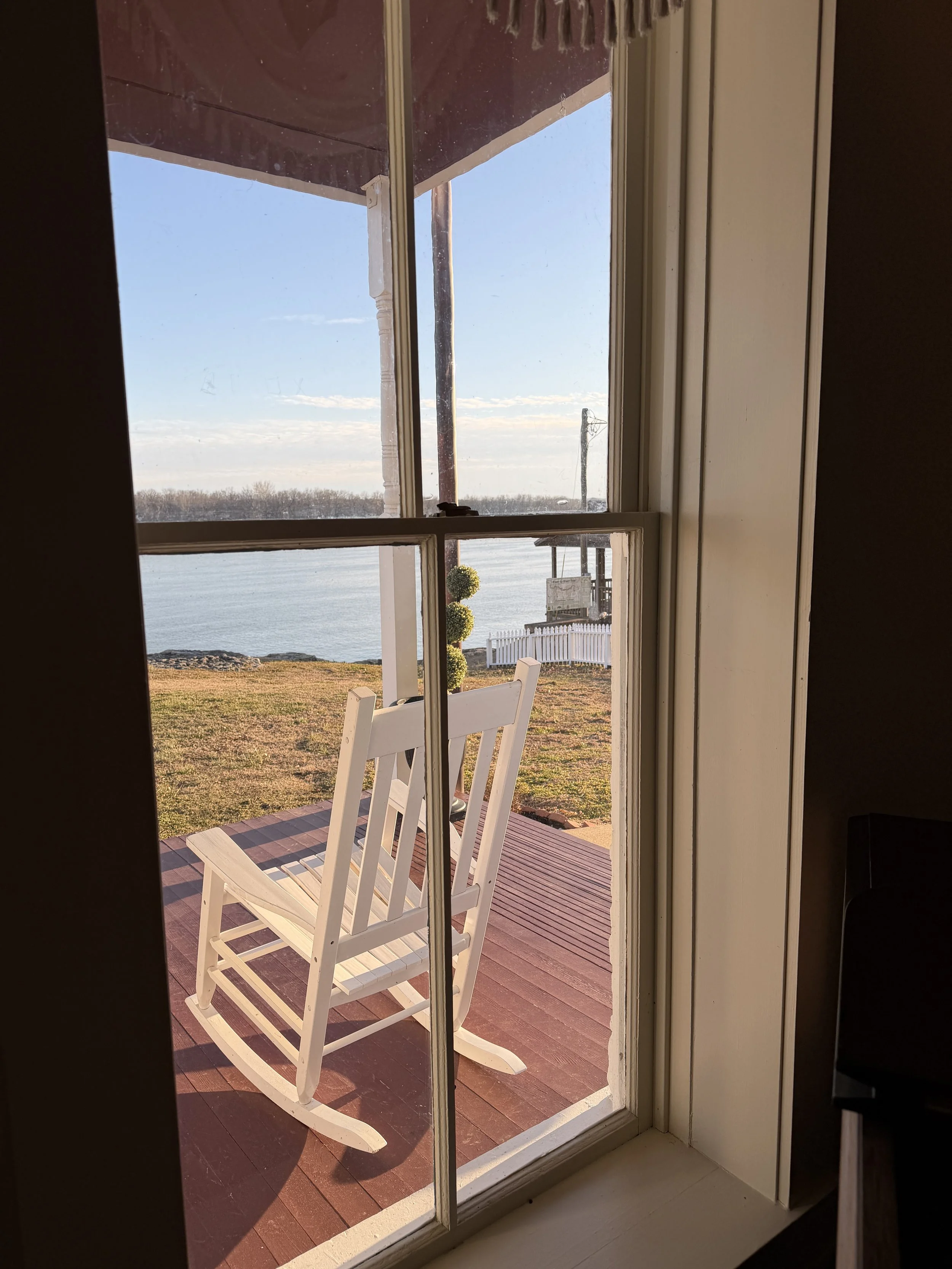 View through a sliding glass door showing a wooden deck with a white rocking chair, overlooking a body of water with trees in the distance and a partly cloudy sky.