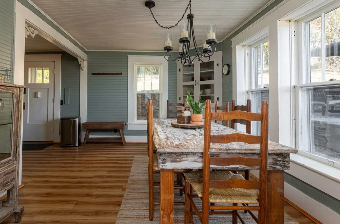 A dining room with a rustic wooden table and chairs, large windows, a chandelier, and light blue walls.