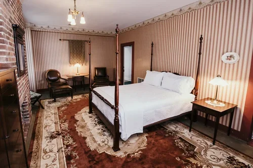 Vintage bedroom with four poster bed, striped wallpaper, area rug, and antique furniture including a side table with lamp, an armchair, and a wooden cabinet.