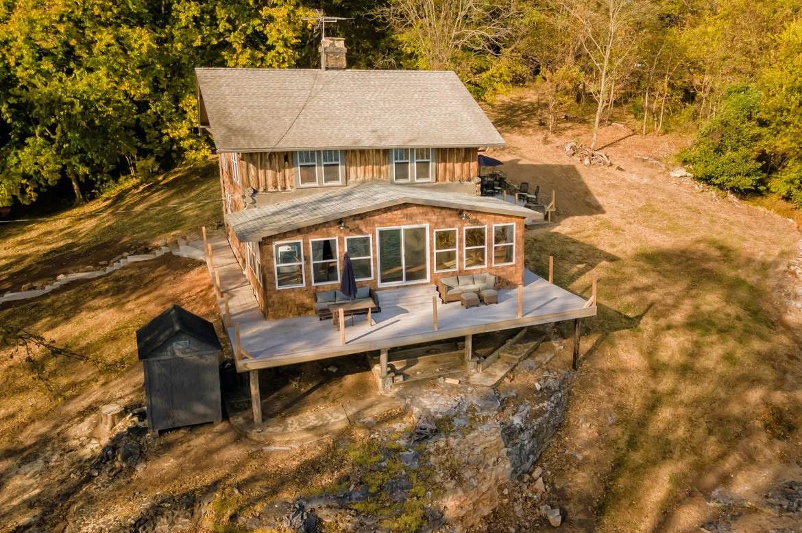 A two-story house built on a rocky hillside with a wooden deck, outdoor seating, and surrounded by trees with autumn foliage.
