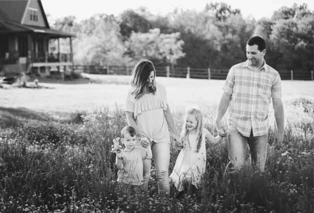 A family of four walking hand in hand through a grassy field with a house and trees in the background, smiling and enjoying the outdoors.