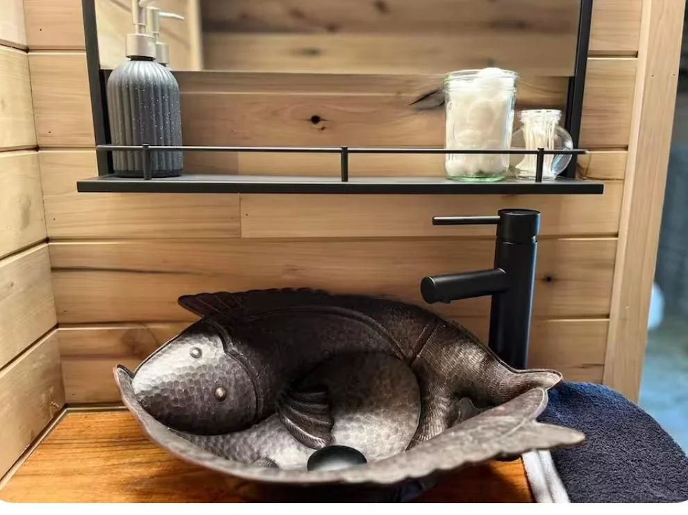 A black faucet above a decorative fish-shaped bowl on a wooden counter, with a metal shelf holding a soap dispenser and glass jars against a wooden wall.
