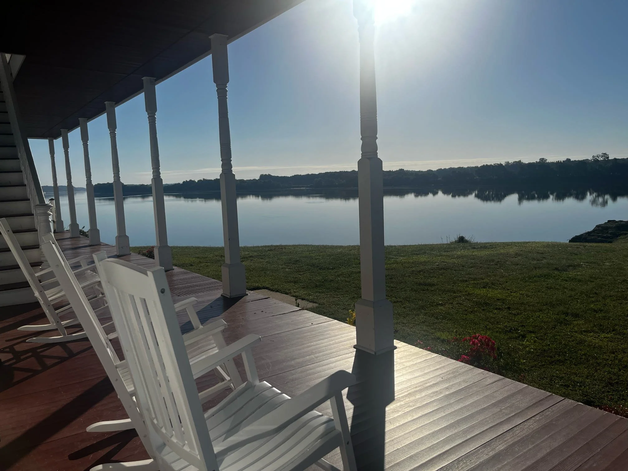 A view from a porch or deck overlooking a large body of water with a tree-covered landmass in the distance. The porch has white chairs, a wooden floor, and white railings, with the sun shining brightly overhead.