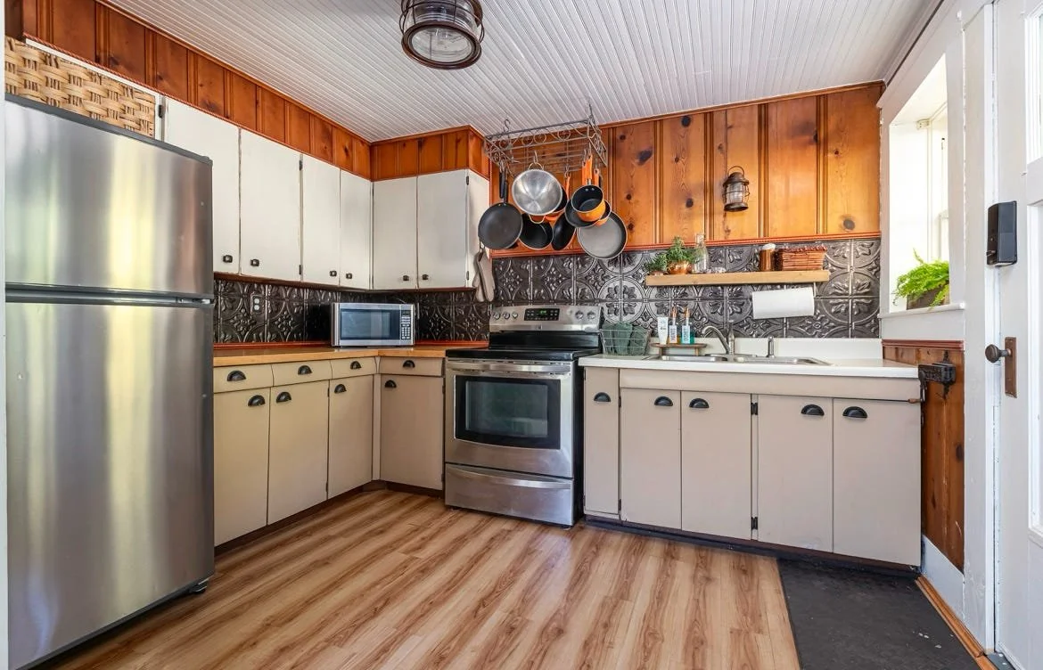 Kitchen with stainless steel refrigerator, microwave, stove, beige cabinet doors, black and brown decorative backsplash, hanging pots and pans, wooden upper and lower cabinets, white countertop, small side window, and wooden accents.