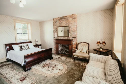 Bedroom with a wooden bed, brick fireplace, upholstered armchair, side tables, and a sofa, with natural light from windows.