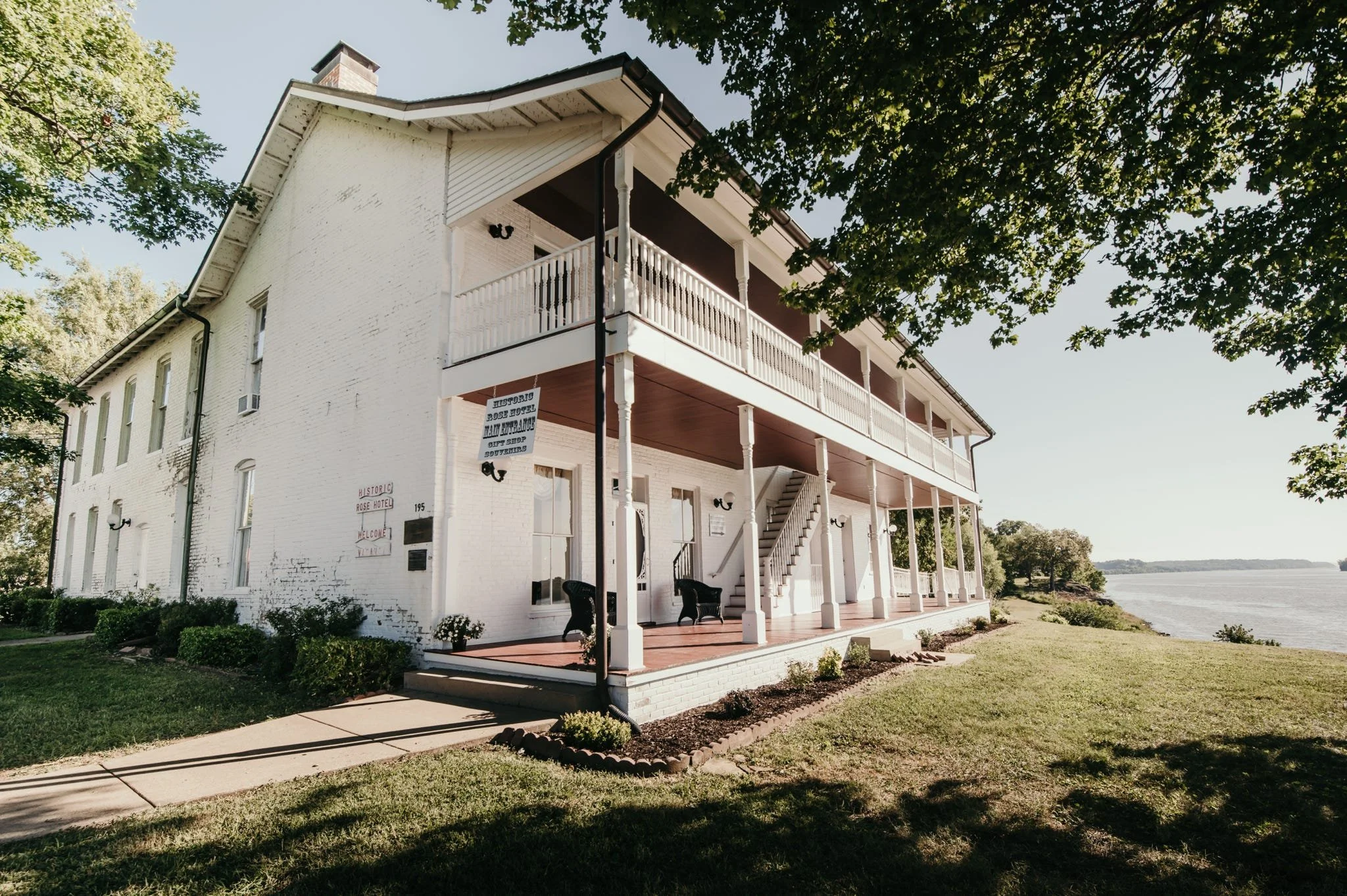 A white multi-story historic hotel building with a large porch overlooking a body of water, surrounded by trees and a manicured lawn.